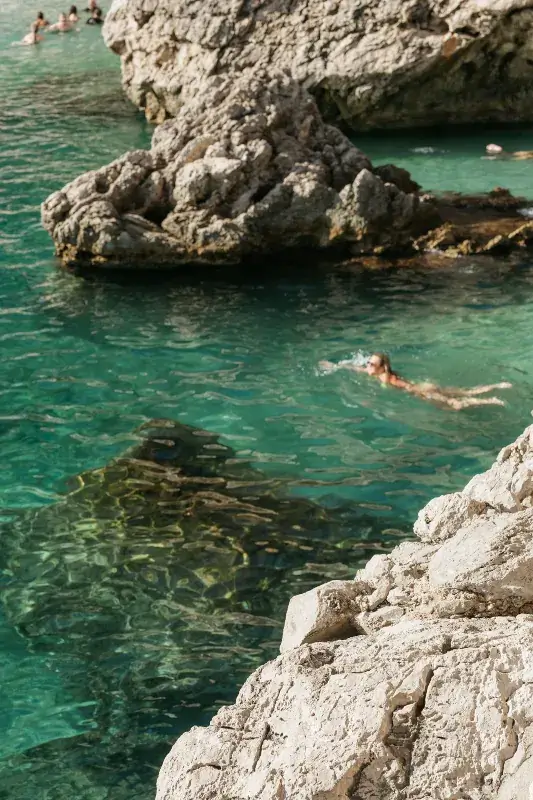 A swimmer enjoying clear turquoise water beside large, rugged rocks. A serene, sunny day; vibrant, inviting coast scene evokes tranquility.