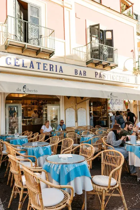 Outdoor seating area of a gelateria with wicker chairs and tables covered in blue tablecloths. People enjoy gelato on a sunny day, conveying a lively and relaxed atmosphere.