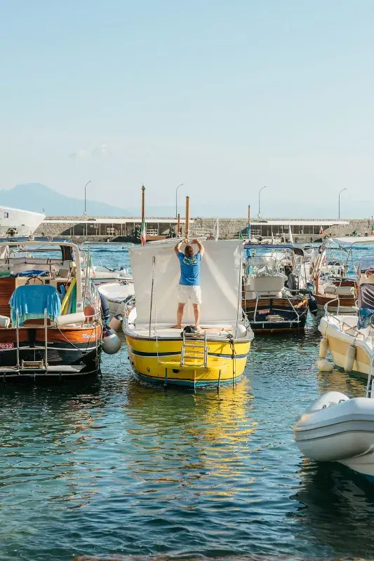 Person in a blue shirt stands on a yellow boat, adjusting a white sail amid a sunny harbor filled with moored boats and calm waters.