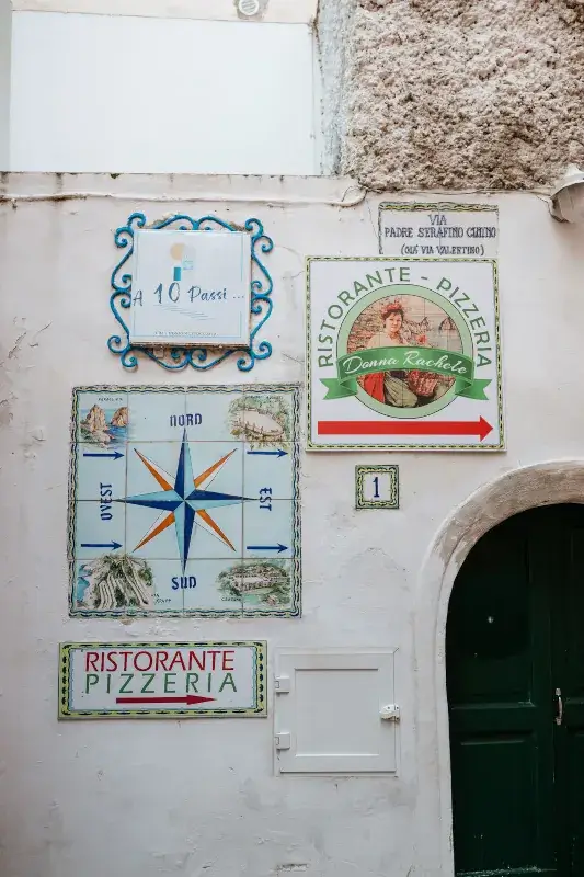 A rustic wall features colorful signs for a pizzeria, compass directions, and a street name, alongside a green arched doorway, evoking a quaint Italian vibe.