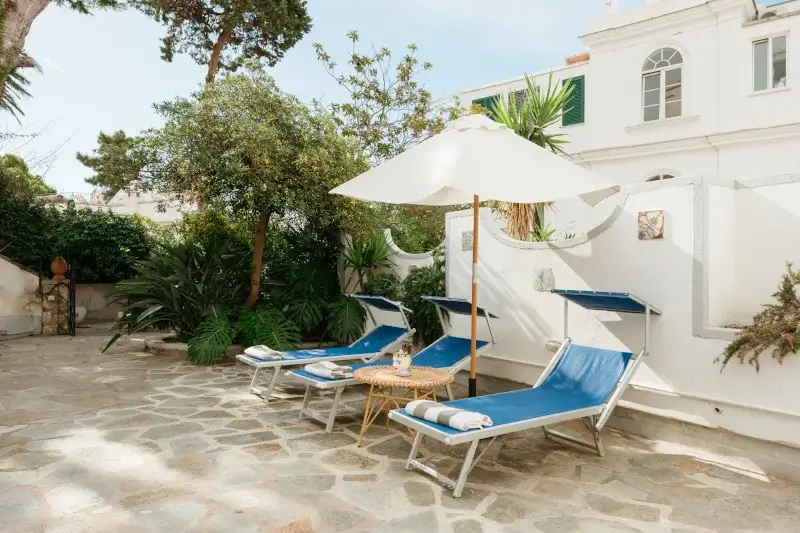 Sunny courtyard with three blue lounge chairs and a white parasol. Surrounded by lush greenery and a white building, creating a serene and inviting atmosphere.