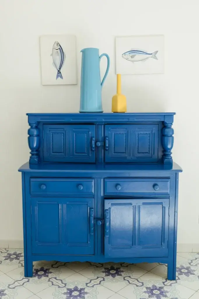 A vibrant blue vintage cabinet with dual, upper and lower cabinets. On top, a tall blue pitcher and a small yellow vase. Two fish paintings are hung above, against a white wall. Decorative tiled floor adds a classic touch.