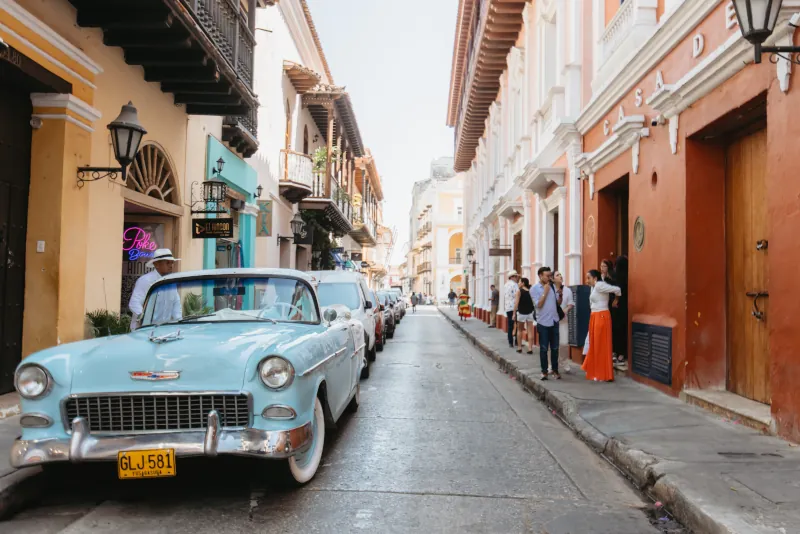 A vintage blue car is parked on a narrow, cobblestone street lined with colorful colonial buildings. People gather and converse on the sidewalk.