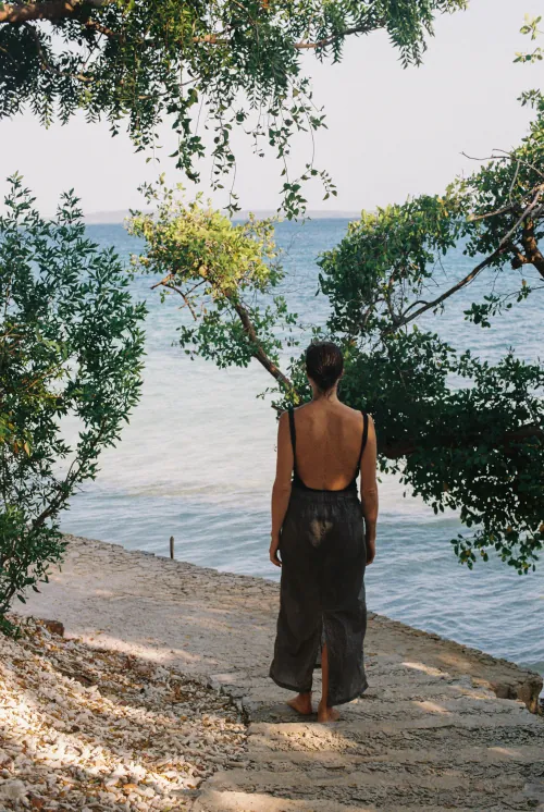 A person in a backless dress walks down a stone path towards a calm, blue sea, framed by lush green trees. The scene conveys tranquility and solitude.