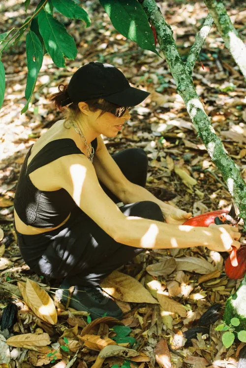 A woman wearing a black cap and sunglasses kneels on a forest floor, surrounded by dried leaves, using pruning shears on a branch. Sunlight filters through the trees.