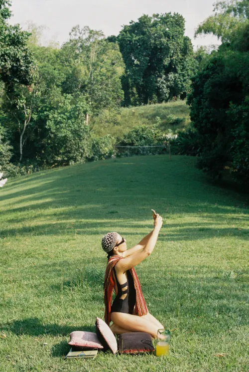 Person sits on grass in a serene park, holding a phone upwards. Surrounding them are cushions, a notebook, and orange juice. Lush trees in the background.