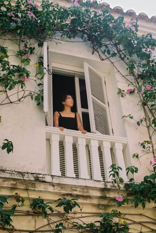 A woman in a black top gazes out from a white balcony framed by pink flowers and green ivy on a sunny day, evoking a sense of calm and contemplation.