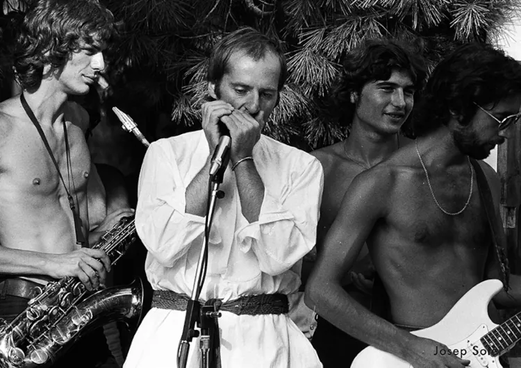 Black and white image of a band performing. A man in a white shirt plays the harmonica, surrounded by shirtless musicians with saxophone and guitar. Energetic and lively.