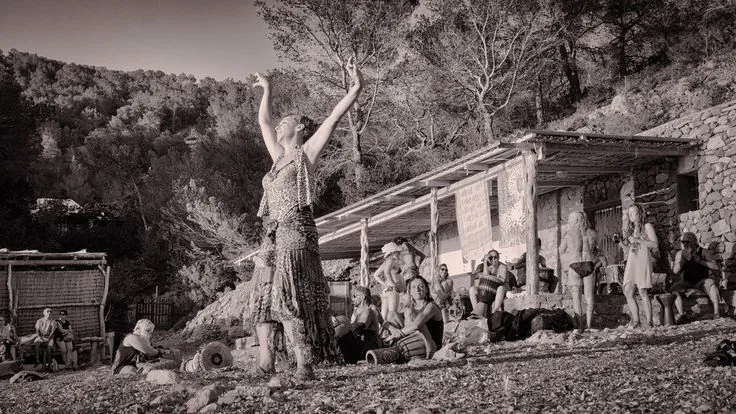 A woman dances with raised arms on a pebble beach, surrounded by an audience and rustic stone buildings. The scene feels relaxed and lively.