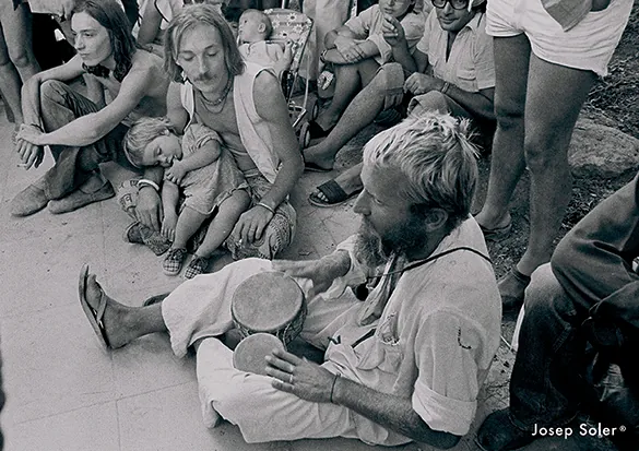Black and white image of a bearded man sitting and playing a drum while a group of people, including children, watch intently. A relaxed, communal atmosphere.