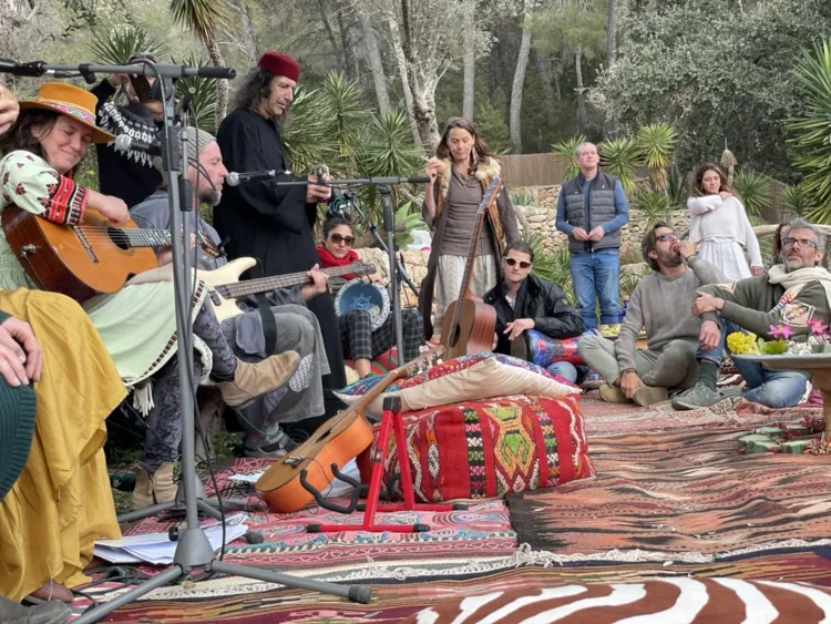 A group of people gathered outdoors, playing guitars and other instruments. They're seated on colorful rugs, surrounded by greenery and trees, creating a relaxed, bohemian atmosphere.