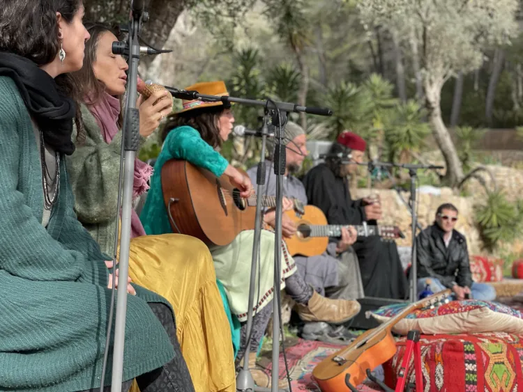 A group of people sit outdoors playing guitars and singing into microphones. They are surrounded by trees and colorful textiles, creating a relaxed, bohemian vibe.