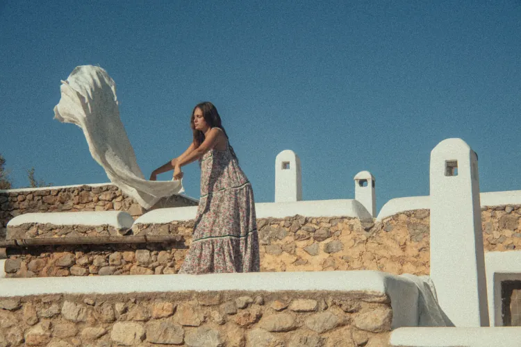 A woman in a floral dress throws a white sheet on a sunlit stone terrace with white walls against a clear blue sky, evoking a serene, breezy atmosphere.
