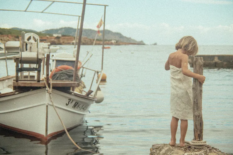 A child in a white towel stands on a dock by a calm sea, holding a post, with a small boat moored nearby. The scene is serene and nostalgic.