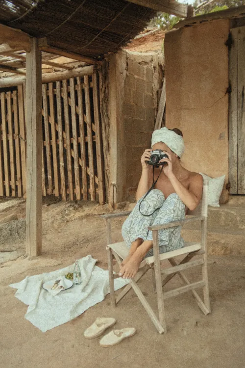 Person sitting in a rustic outdoor setting, wrapped in a towel turban, casually taking a photo with a vintage camera. Relaxed, serene atmosphere.