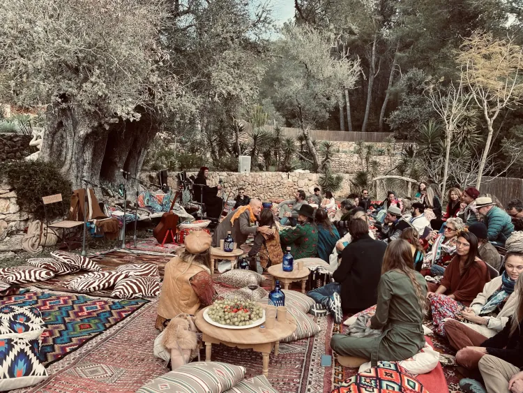 Outdoor gathering with people seated on colorful rugs and cushions amidst nature. Tables hold fruit and drinks, creating a relaxed, communal atmosphere.