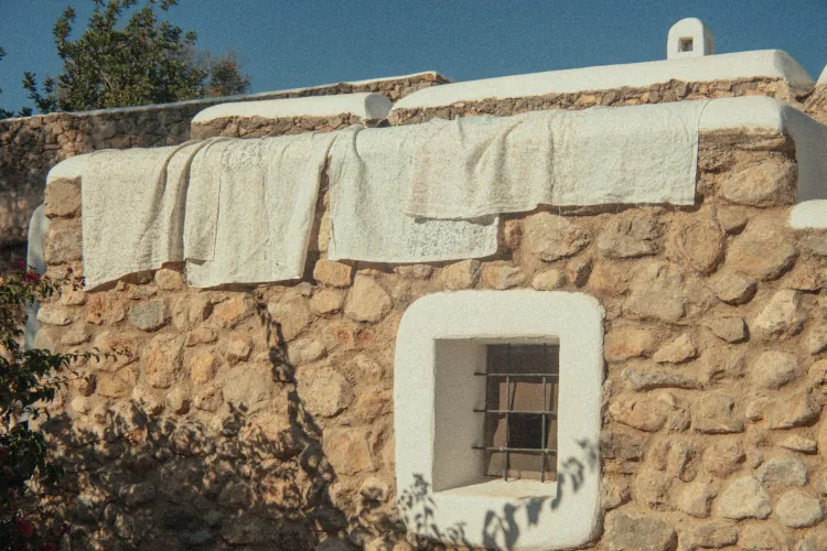 Stone house with whitewashed accents under a clear blue sky. Towels hang over the edge, casting shadows. Rustic, serene Mediterranean vibe.