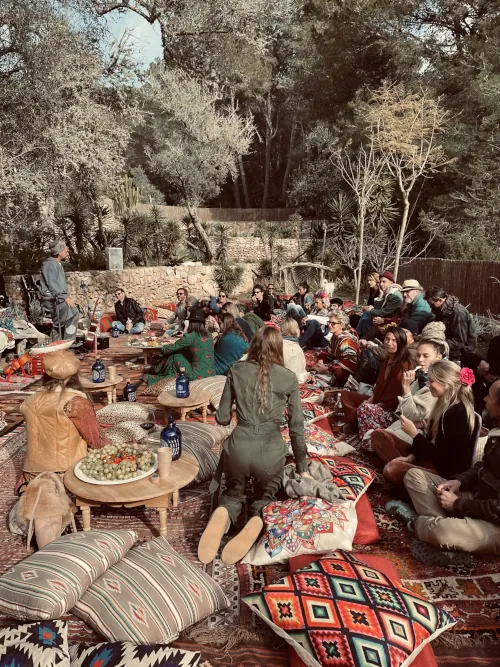 A diverse group of people is seated outdoors on vibrant, patterned cushions, surrounded by trees. The scene is relaxed and communal, with a bohemian vibe.
