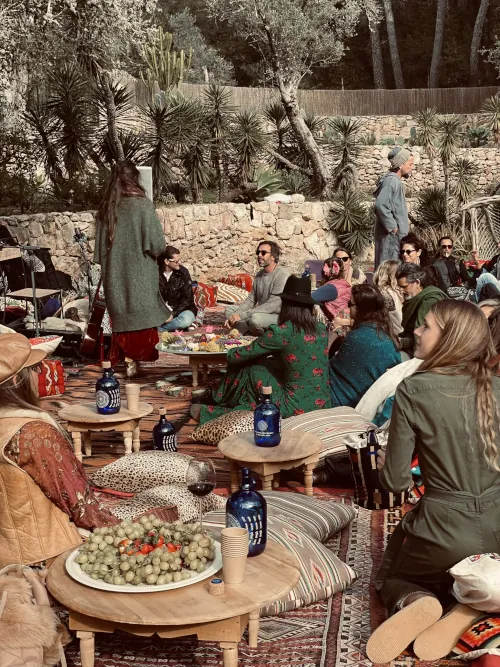 A relaxed outdoor gathering in a rustic garden with people sitting on cushions around wooden tables. Plates of food and drinks are shared, creating a cozy, bohemian vibe.