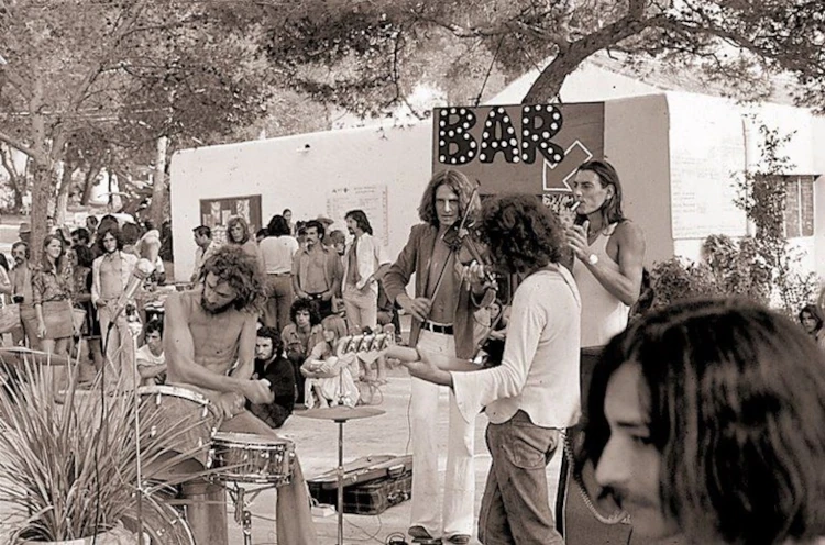 Black and white image of an outdoor concert with four musicians playing instruments near a bar sign. A relaxed crowd listens under trees. 1970s vibe.