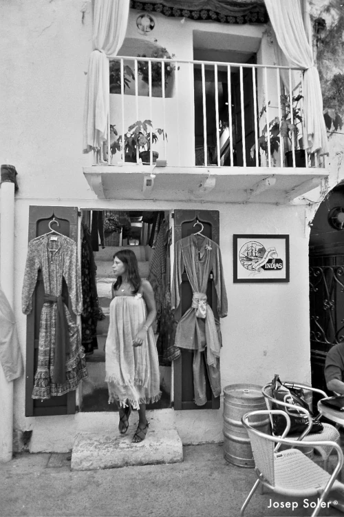 Black and white photo of a woman in a dress standing outside a shop. Dresses hang on either side of her. An upstairs balcony with plants is visible.
