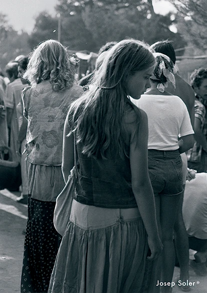 Black and white photo of a young woman with long hair in a flowing skirt, walking through a crowded outdoor scene, evoking a nostalgic and peaceful mood.