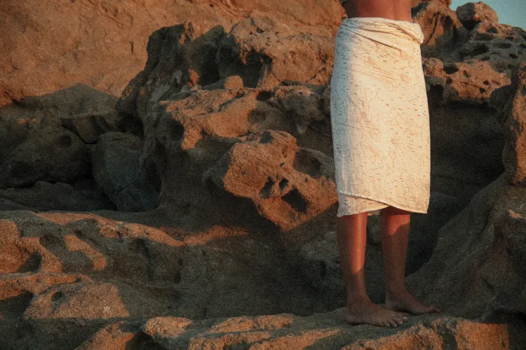 Person wrapped in a white towel standing barefoot on rugged, sunlit rocks. The scene conveys a sense of calm and natural beauty.