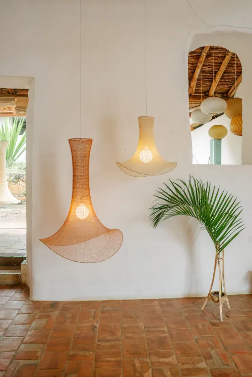 Hanging woven lamps in a rustic room with white walls, terracotta flooring, and a potted palm. A doorway shows greenery outside, adding warmth.