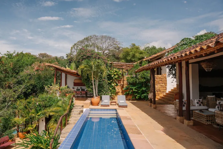 Luxurious villa patio with a long, narrow pool flanked by sun loungers, tropical plants, and rustic-style architecture under a clear blue sky. Relaxing ambiance.