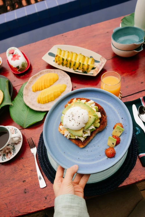 A hand holds a blue plate with avocado toast topped with a poached egg. Surrounding are plates of sliced mango, pineapple, and a cup of coffee on a wooden table.
