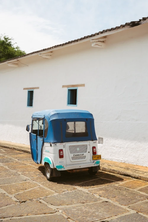 A blue and white tuk-tuk parked on a stone path against a white wall with blue windows, under a sunny sky, evoking a quaint, serene atmosphere.