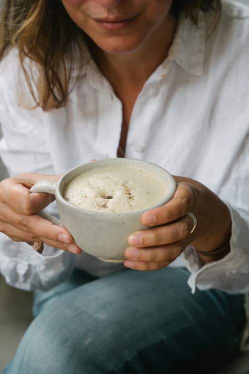 A person in a white shirt holds a mug of frothy coffee, sitting casually. The scene conveys warmth and relaxation, with a cozy and inviting atmosphere.