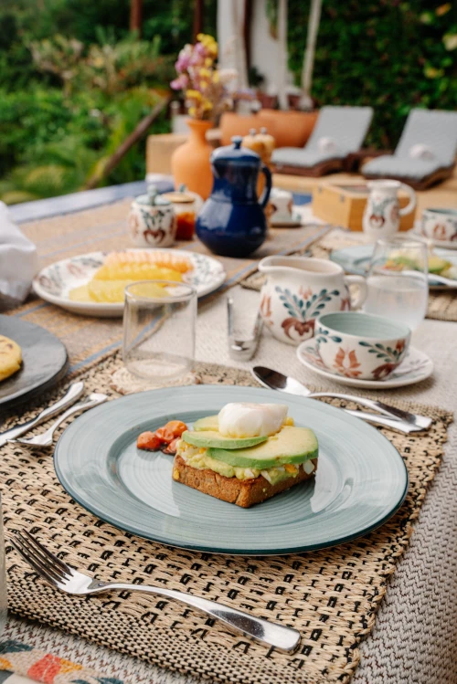 A breakfast table set outdoors features avocado toast topped with a poached egg on a blue plate. Floral teacups, a blue teapot, and lush greenery in the background create a serene, inviting atmosphere.