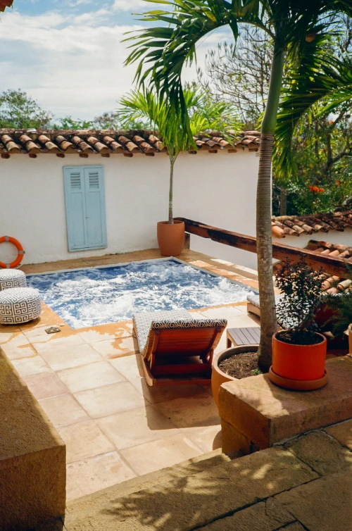 A cozy courtyard features a small jacuzzi surrounded by terracotta tiles, with a sunbed and tropical plants. A rustic white wall with a light blue door adds charm.