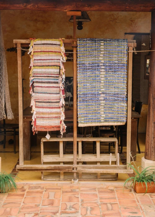 Two vibrant woven rugs hang on a wooden loom in a rustic setting. The left rug features warm colors, while the right rug is in cool blue tones.