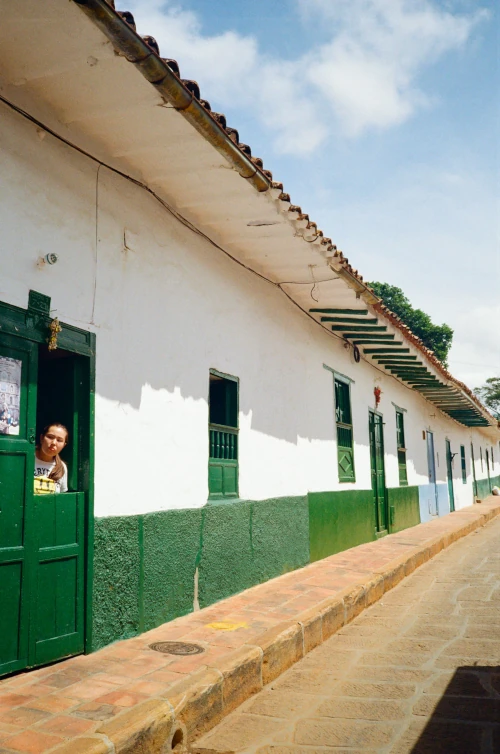A person peeks out from a vibrant green doorway of a long, white colonial-style building on a sunny day. The scene conveys a calm, quaint atmosphere.