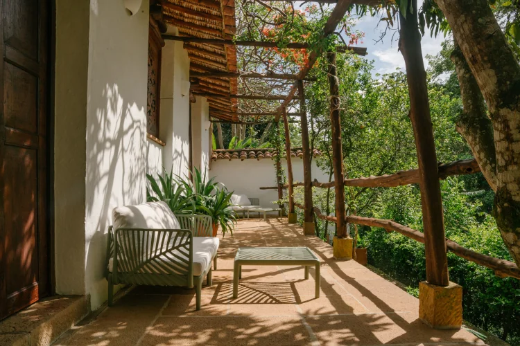 Sunny outdoor patio with white cushioned chairs, wooden beams, and lush greenery. Dappled sunlight creates a peaceful, inviting atmosphere.