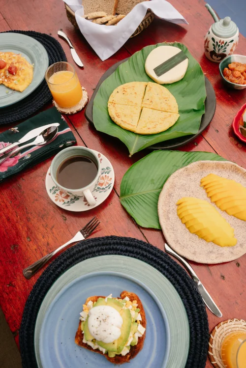 A vibrant breakfast spread on a wooden table, featuring sliced mango, cheese arepas on leaves, avocado toast, a cup of coffee, and a glass of orange juice.