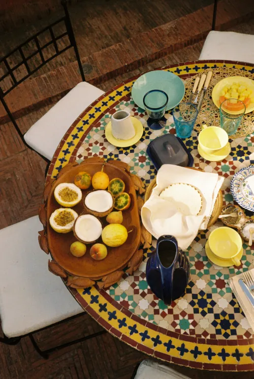 A colorful mosaic table displays a vibrant breakfast setting with cut fruits, various cups, and plates, evoking a lively and inviting atmosphere.