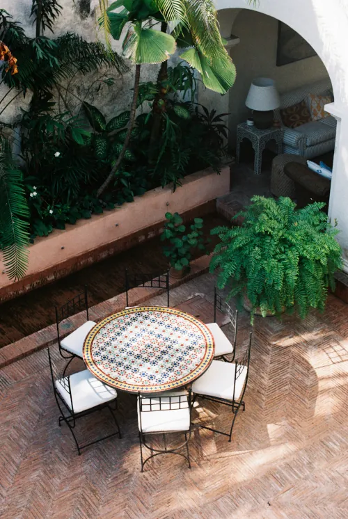 Sunny courtyard with a round, mosaic-patterned table and six chairs. Lush greenery, including ferns and palm plants, surrounds the space. Cozy, inviting atmosphere.