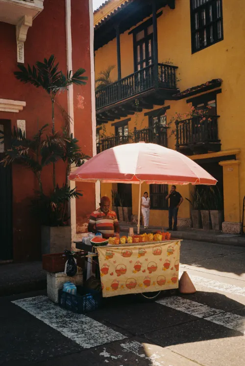 Street vendor with a colorful cart under a pink umbrella sells fruit on a sunny day, set against vibrant red and yellow colonial buildings.