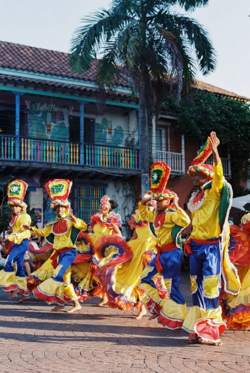 A vibrant group of dancers in colorful, traditional costumes perform energetically in a sunny, cobblestone street. Palm trees and festive buildings line the background.