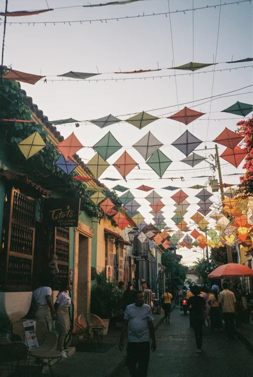 Colorful kites hang over a lively, narrow street lined with vibrant buildings. People stroll below, creating a festive and cheerful atmosphere.