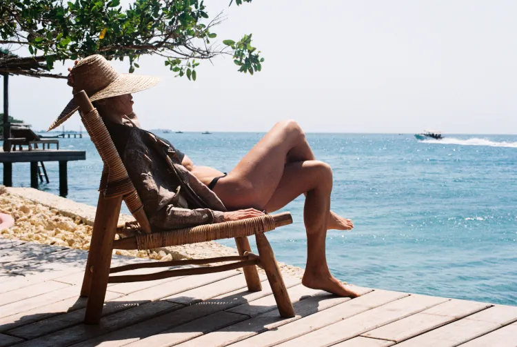 A person relaxes on a wooden deck chair by the ocean, wearing a wide-brimmed hat and a shirt. Sunlight highlights the calm, serene beach setting.