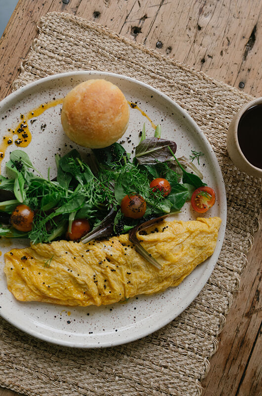 A gourmet breakfast at a luxury villa in Barichara, featuring a fresh omelet with cherry tomatoes, a side salad, and a traditional roll on a rustic wooden table.