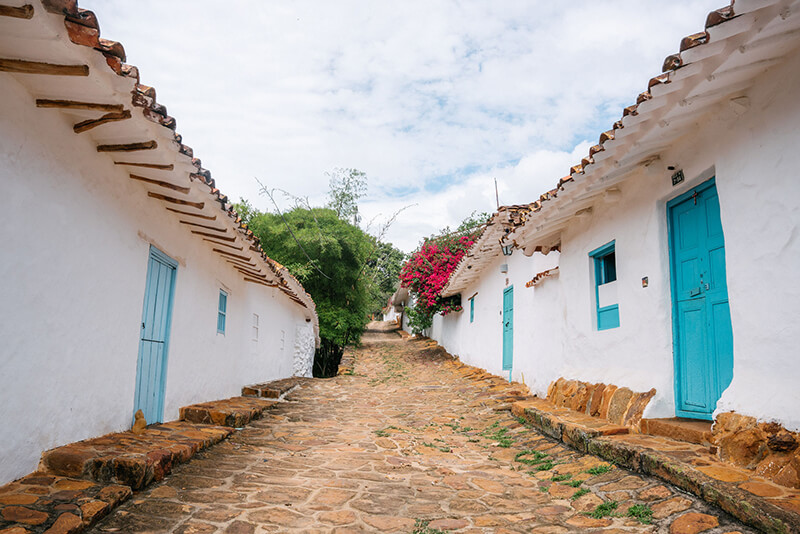 Narrow cobblestone street in Barichara, Colombia, lined with traditional white colonial houses featuring bright blue wooden doors and red bougainvillea.