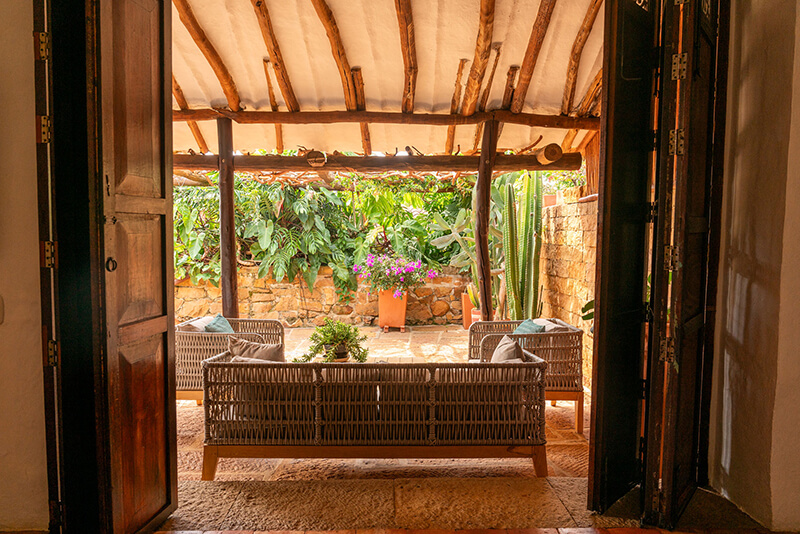 View through tall wooden doors of a luxury villa in Barichara, showing a covered stone patio with modern woven lounge furniture and a lush tropical garden.