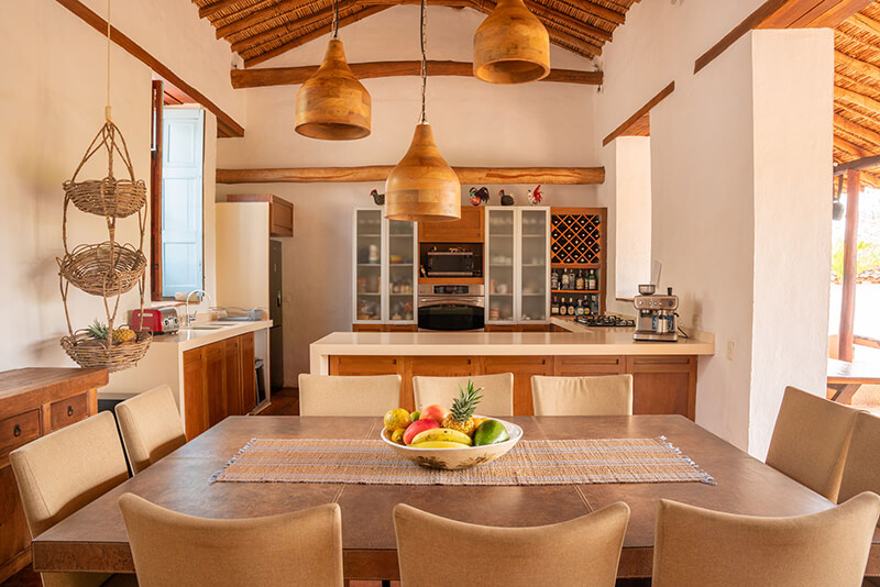 Open plan kitchen and dining area in a luxury villa in Barichara, featuring a large wooden table, pendant lights, and rustic high ceilings.