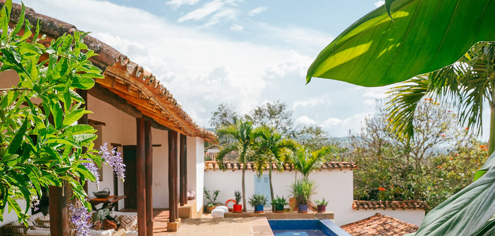 Wide-angle view of a luxury villa terrace in Barichara featuring a blue plunge pool, wooden pillars, and lush tropical greenery under a bright sky.