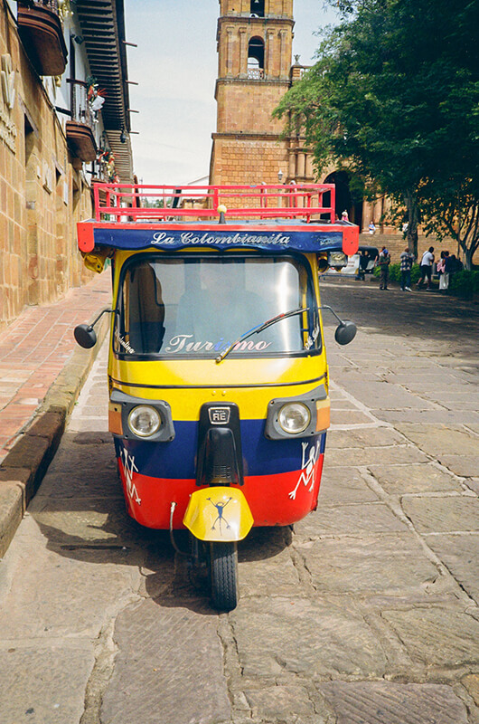 A colorful yellow, blue, and red tuk-tuk parked on a cobblestone street in Barichara, Colombia, with the colonial sandstone cathedral in the background.